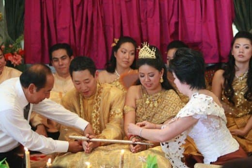 The couple kneels while holding a sheathed sword as their guests bestow blessings by tying a red string around each of their wrists.