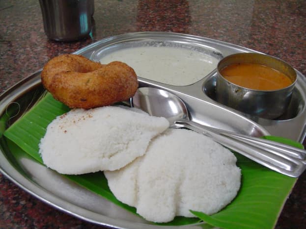 The idli is in white. The brown colored round dish is vada which is also many times served with idli sambhar if asked for it. Vada is made by frying a batter of dal, lentil, gram flour or potato. They have a shape similar to a doughnut.