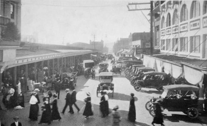 Scene in Pike Place Market c. 1915. Looking roughly northwest on Pike Place. At left, Main Market (later rebuilt as Main Arcade). At right, Corner Market and Sanitary Market. In distance, Washington National Guard Armory.