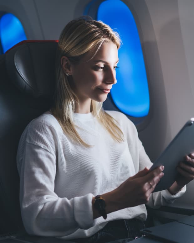 A woman sits in her airplane seat, looking at a tablet in front of a bright window. The other electrochromic windows behind her are dark and shaded.
