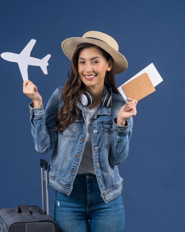 Brunette woman in denim and straw hat holding a paper airplane outline and passport/ticket, with luggage next to here