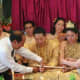 The couple kneels while holding a sheathed sword as their guests bestow blessings by tying a red string around each of their wrists.