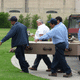 The hearse has brought the casket to the grave site and the crew is carrying the casket to the grave to place it in the liner.