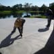 The upright worker has a hopper filled with the CoolDeck solution and he's spraying it on the concrete. The worker who's leaning down has a trowel to spread and flatten it. He's wearing spikes strapped on his shoes to avoid treading on the wet glop.