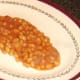 Baked beans are plated as a bed for sausage roll dog