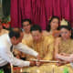 The couple kneels while holding a sheathed sword as their guests bestow blessings by tying a red string around each of their wrists.