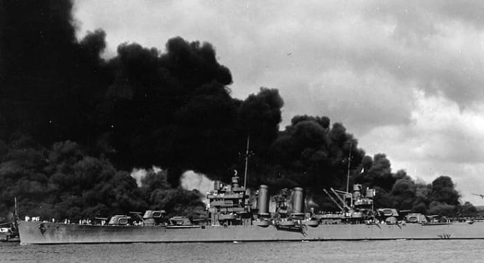 The U.S. Navy light cruiser USS Phoenix (CL-46) steams down the channel off Ford Island's "Battleship Row", past the sunken and burning USS West Virginia (BB-48), at left, and USS Arizona (BB-39), at right, 7 December 1941.