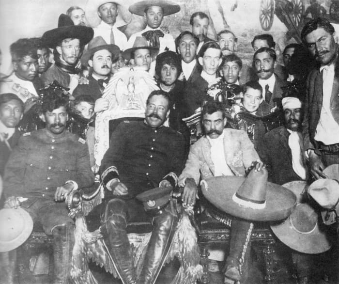 Pancho Villa (in the Presidential chair) chats with Emiliano Zapata at Mexico City. Tómas Urbina is seated at far left, Otilio Montaño (with his head bandaged) is seated to the far right.