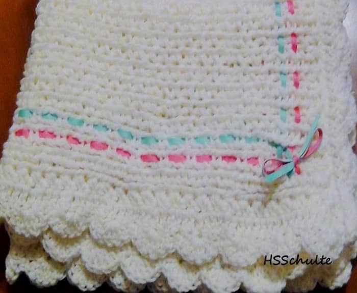 The Knifty Knitter Infant Blanket is 3.5 feet x 2 feet. It is loomed with an 8 wrap cast-on, the fashion stitch, and the crochet cast-off. The shell stitch is crocheted around the edges and a ribbon is woven through the knit.