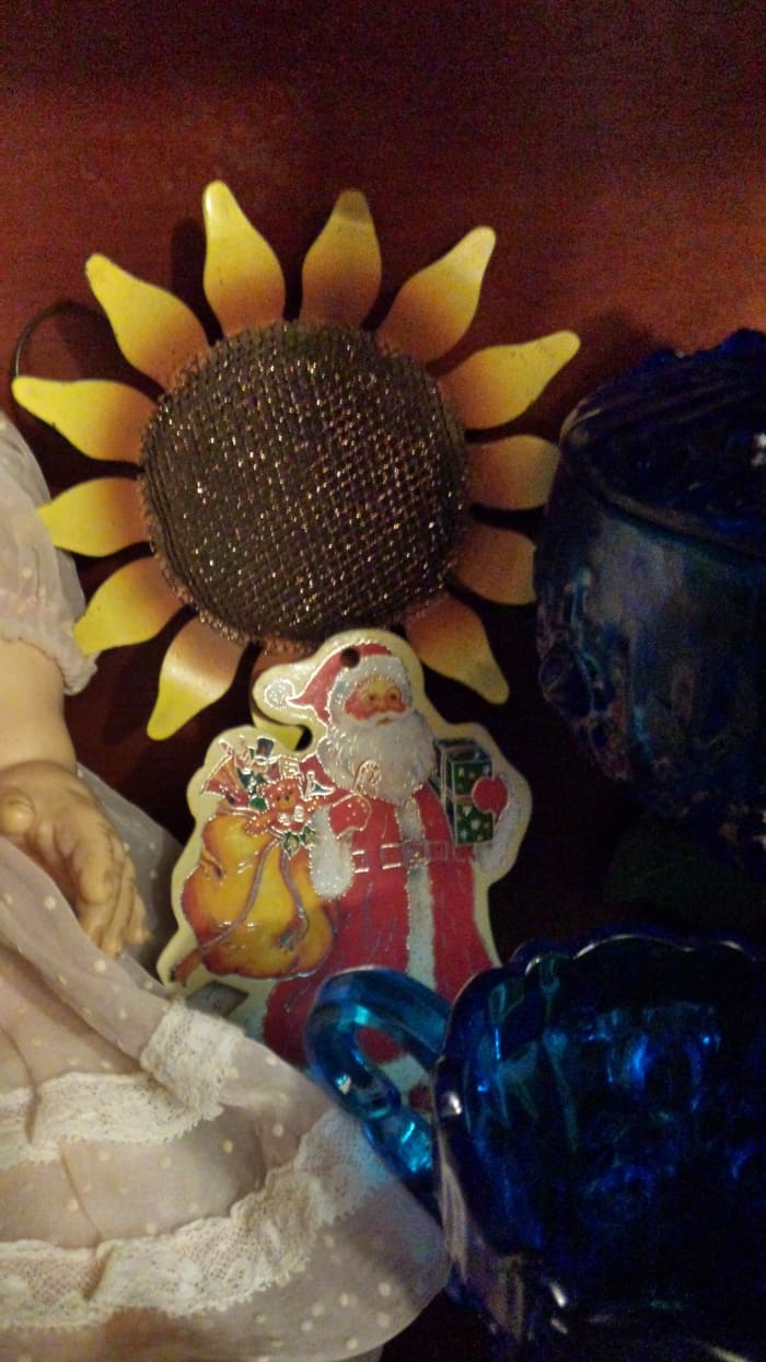 Photograph of vintage cardboard cutout Santa ornament with gold outlines. He stands in front of a metal sunflower and beside antique blue glass. There is a baby doll seen only in part on the photo's left side.