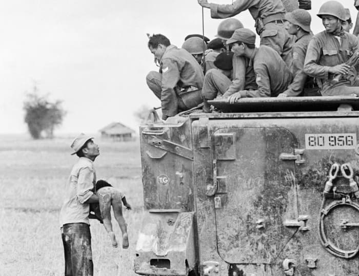 A distraught father holds the body of his child as South Vietnamese Rangers look down from their armored vehicle, March 19, 1964. The child was killed as government forces pursued guerrillas into a village near the Cambodian border.