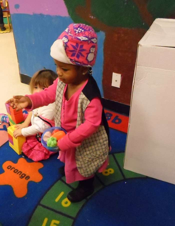 One little girl on the floor playing, and another girl standing with dress up close.