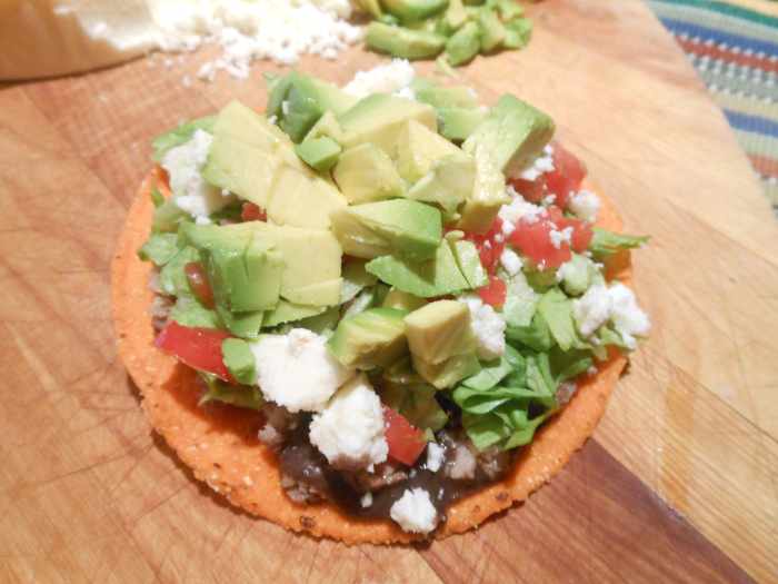 A beef tostada includes black beans, thinly sliced steak, shredded lettuce, chopped tomato, crumbled queso fresco, and chunks of avocado.
