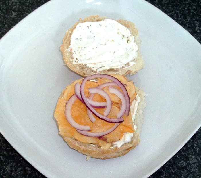 Herb and spice cream cheese is spread on bread roll before cheese and onion crisps and red onion slices are added