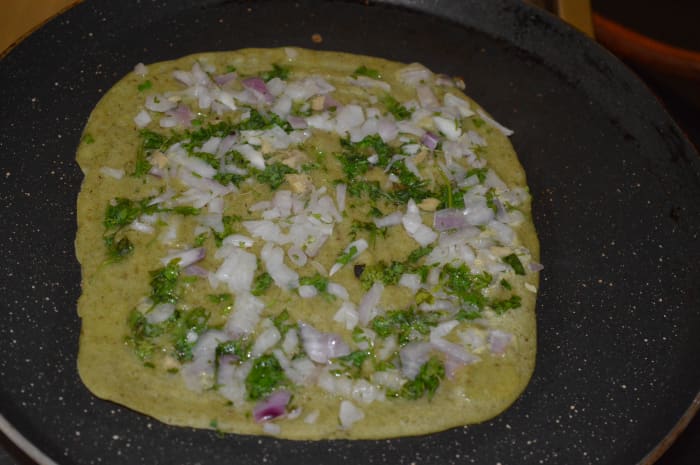 Step five: Heat a pan. Grease it with oil or ghee. Add 1 1/2 ladle of batter in the center. Spread it to make a square or round shape. Spread a teaspoon topping mix on the top. Add a few drops of oil or ghee on the top. Press the topping down a bit.
