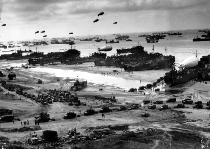 WW2: Normandy Invasion, June 1944. Landing ships (LSTs) putting cargo ashore on one of the invasion beaches, at low tide. Note the barrage balloons overhead and Army "half-track" convoy forming up on the beach.