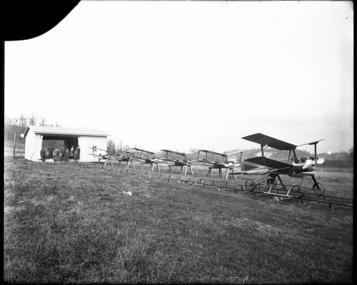WW1: A Kettering Bug on the rail cart ready to be launched. Five other Bugs are lined up alongside the rail track resting on sawhorses. The rail track runs from a small building with Dayton-Wright employees standing in the opening observing the Bugs