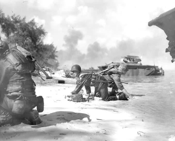 WWII: Saipan beachhead. Two Marines keep down low while they crawl to their positions as Japanese fire whines overhead. Their landing craft was hit by a Japanese mortar. In the background are Buffalos which supported the Marines in their landing.