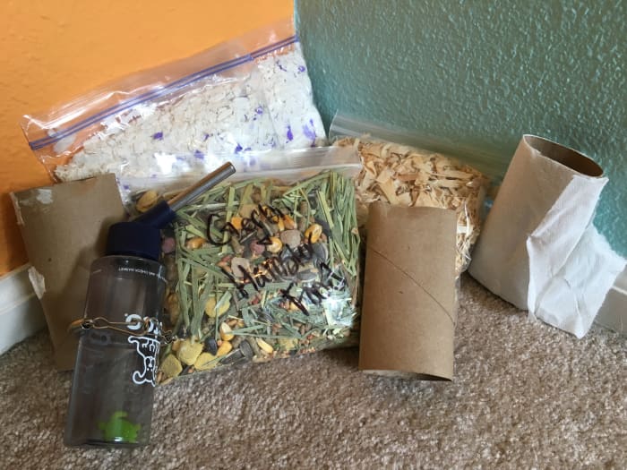 Example of supplies for a hamster or gerbil. Pictured are two bags of different types of bedding, water bottle, chewable toilet paper rolls, and a bag of food mixed with Timothy hay. Travel cage is the only other thing needed.