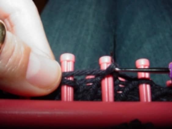 Knitting off no-wrap stitch on a Knitfy Knitter loom. This is a photo of the no-wrap stitch in progress. In the picture, the hook is being used to knit off the pink loom, as the yarn is stretched just above it across the peg. I hold the working yarn.
