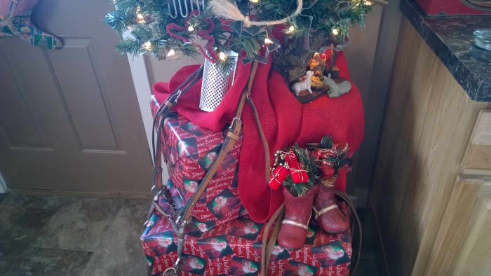 Large empty boxes wrapped with Christmas paper form a stand for a small tree which has been decorated with items like cookie cutters, bits of rope, an antique horse bridal, and a couple of red boot shaped kitchen utensil holders filled with garland.