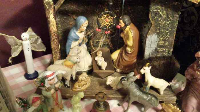Antique creche featuring Mary, Joseph, and baby Jesus alongside sheep, shepherds, a donkey, and a single wiseman. A snowman angel sits to one side, there is a dreamcatcher over the baby, and a Christmas bell in front of the scene.