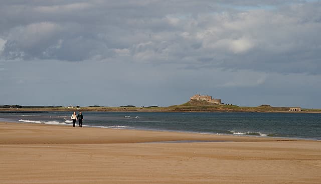 A couple walk along the shoreline with Lindisfarne Castle on Holy Island in the background.