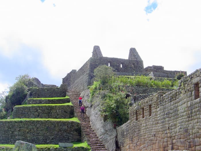 Travelers enjoy descending from an upper level down the stone steps along walls, all constructed by Incas prior to 1500 A.D., while viewing the architecture, with vivid grass and lush shrubbery adding green to the surroundings.
