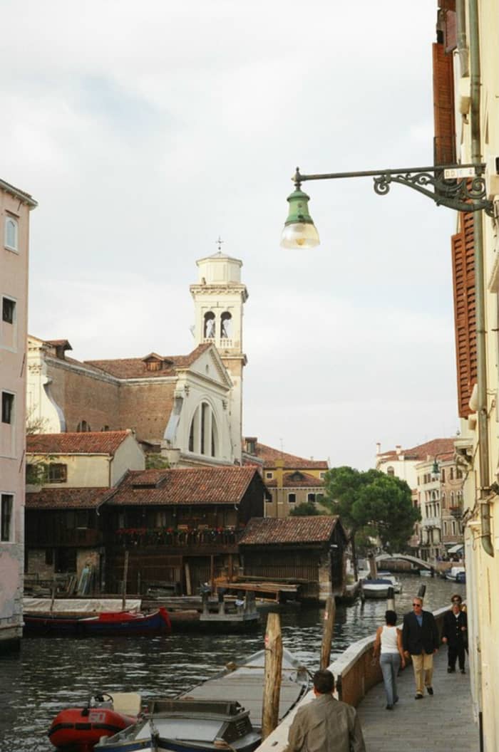 Pedestrians walk alongside the canal near the Squero di San Trovaso boatyard, where gondolas are made, with the Church of San Trovaso in the background.