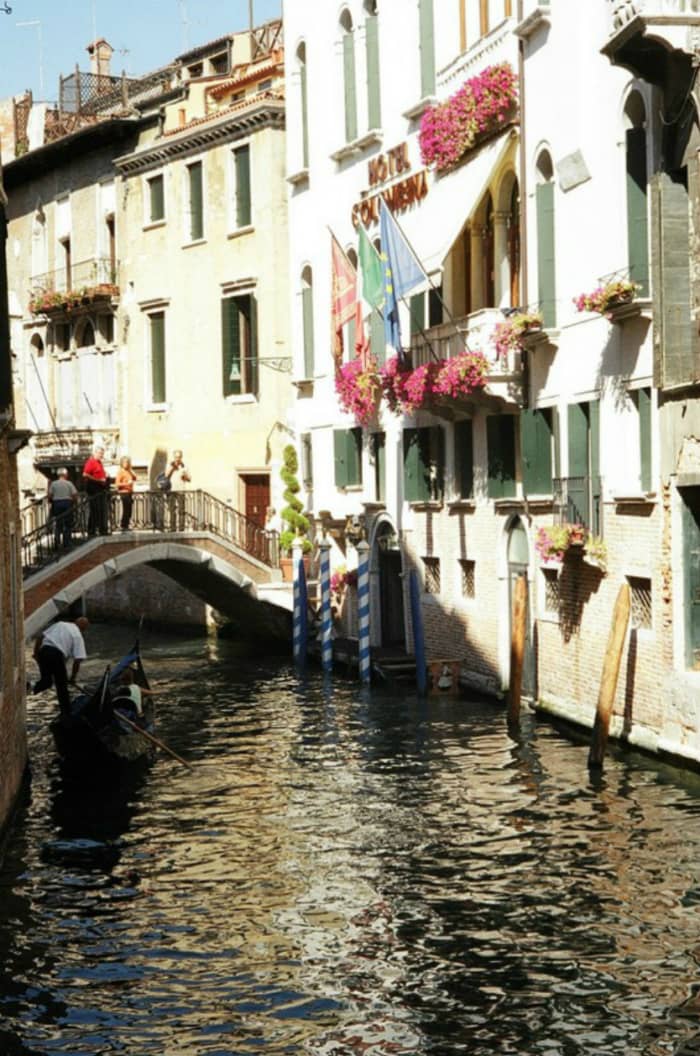 Flowers and flags adorn a hotel overlooking a bridge over a small canal, with a gondola passing through.