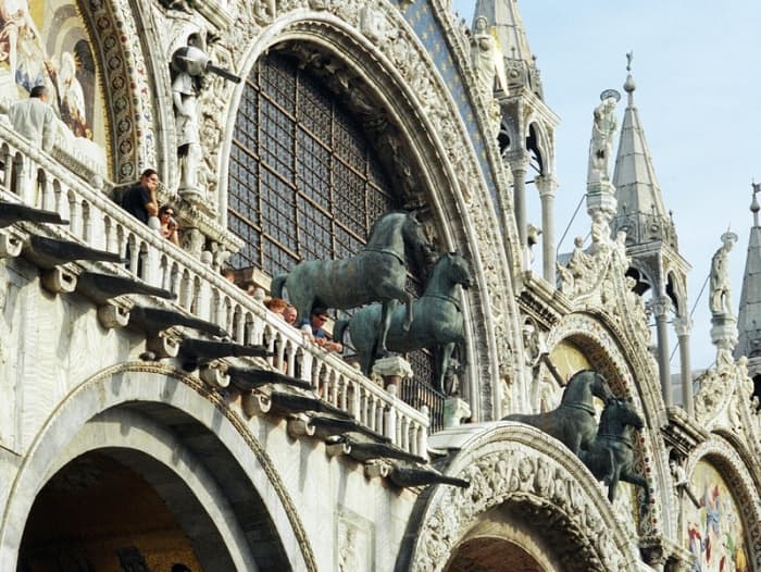 A close-up of the archways of the ornate facade of St. Mark's Basilica, with the replicas of the ancient bronze horses on the balcony, accompanied by tourists. The original horses were placed inside the Basilica to protect them from the weather.