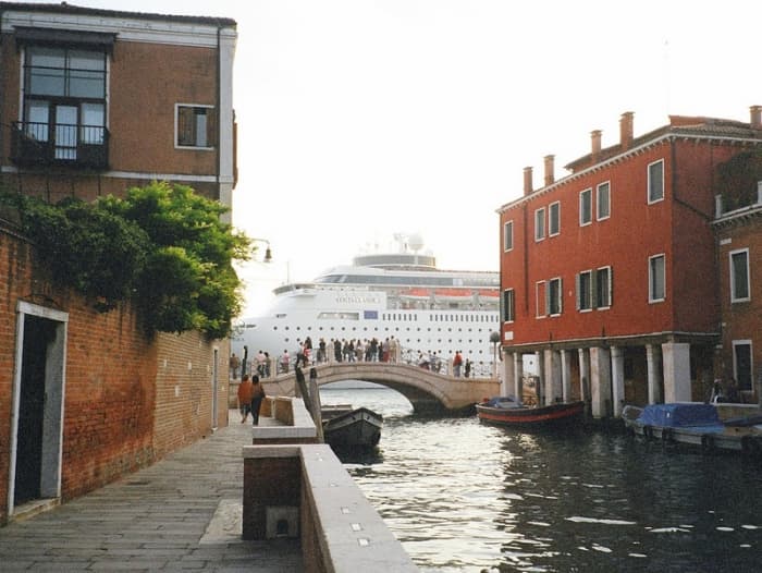 A large cruise ship passes, dominating the view, as tourists look on from a bridge over a canal in Venice.