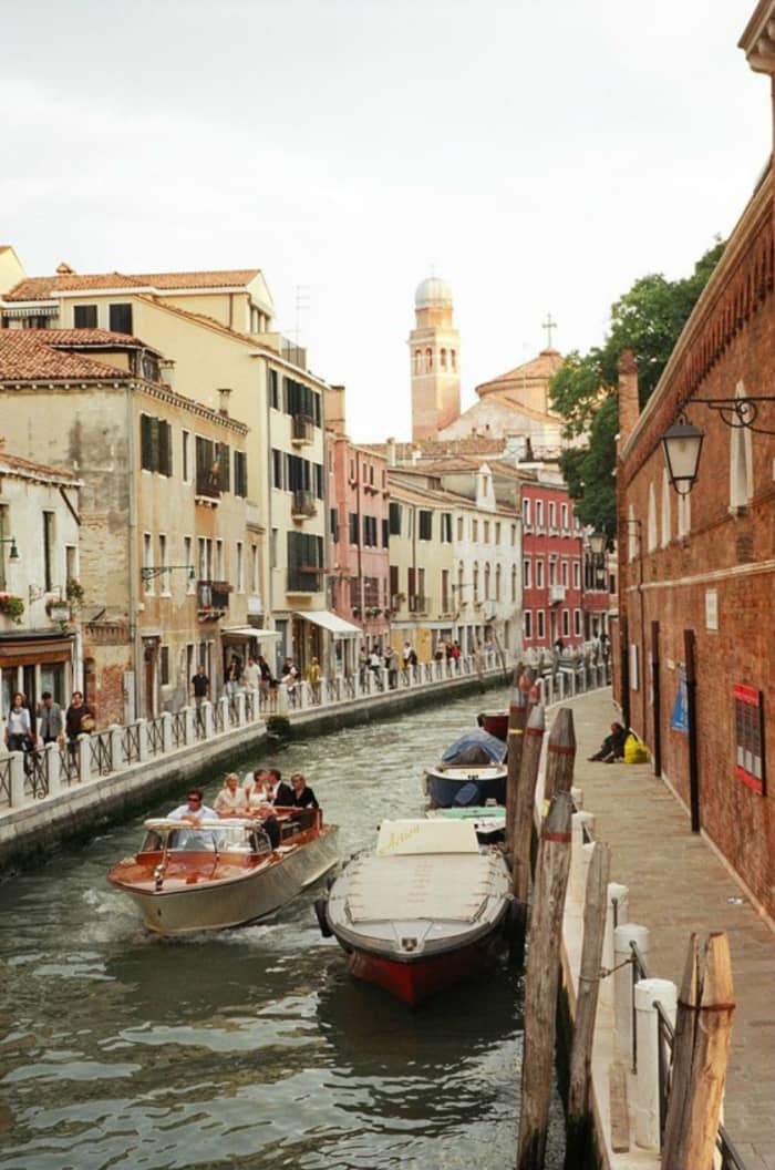 A water taxi in the mode of a classic speedboat carries a wedding party down a canal in Venice, flanked by walkways of sightseers celebrating the beauty of this unique city, where, instead of streets, there are only waterways.