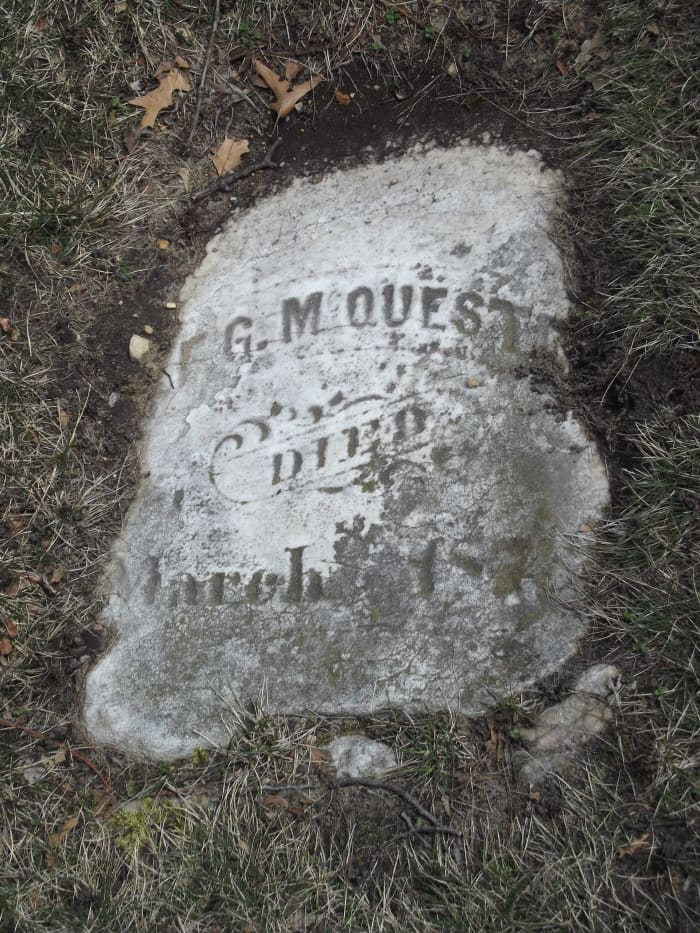 A somewhat square tombstone flat on the ground that is mostly intact. The text is relatively faded. The grass has grown over the sides. Evergreen Cemetery, Muskegon, MI.