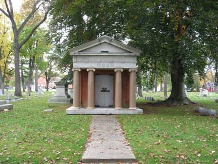 Image of mausoleum with columns out front and grey granite doors. Mausoleum itself is sandstone colored with columns of matching terracotta granite. Moon family crypt, Evergreen Cemetery, Muskegon, MI.