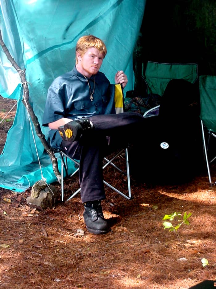 Young man in front of a tarp while camping. If it rains, you need covered space for eating or just hanging out.(Sorry to interrupt your reading, son)