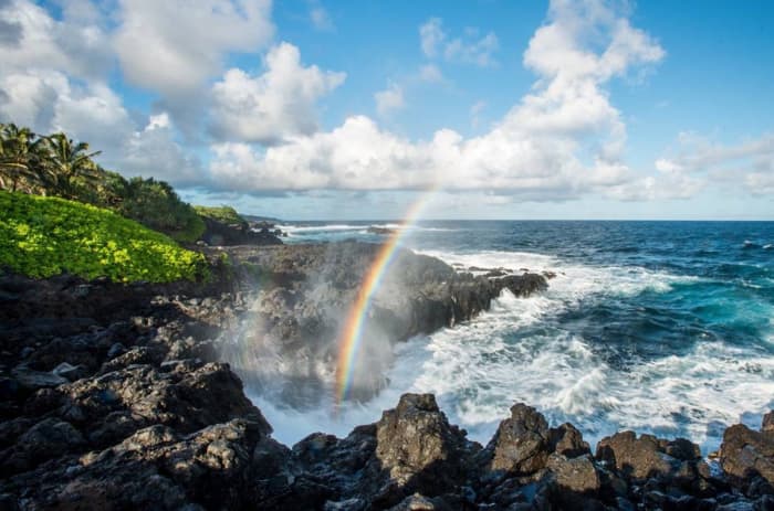 A rainbow forms in the mist of the ocean crashing on a rocky shore, in the Kipahulu area of Haleakala National Park, Maui, HI