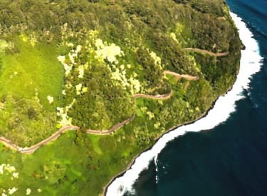 An aerial view of the Hana Highway gliding 68 miles along the shore of Maui, with beautiful views of the surf below, crossing many bridges over creeks headed for the sea.