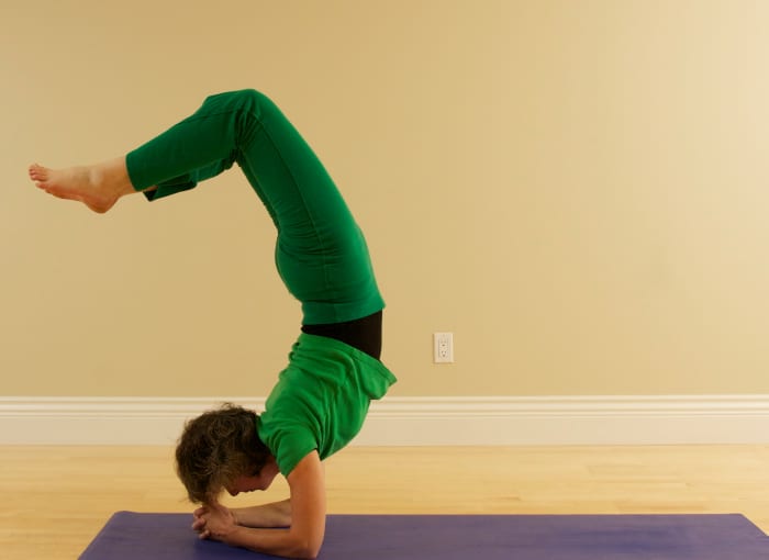 For Vrischikasana Scorpion from Headstand, bend the knees and drop the feet toward the back body as you lift the head and look forward. To come out, plant the head in the hands again first, then straighten the legs.