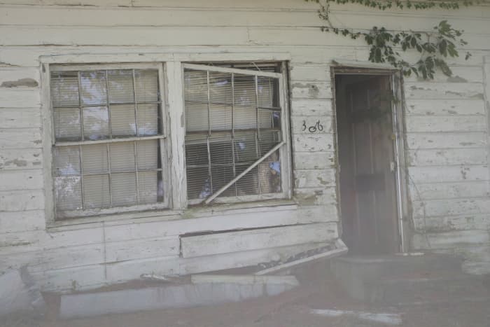 Mist flows from the doorway of an abandoned house, while something appears to peek from behind the window.