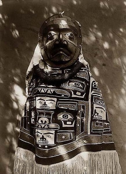 From Alaska, this is a woman in a fringed Chilkat blanket, a Hamatsa neck-ring and a mask representing a deceased relative who had been a shaman.