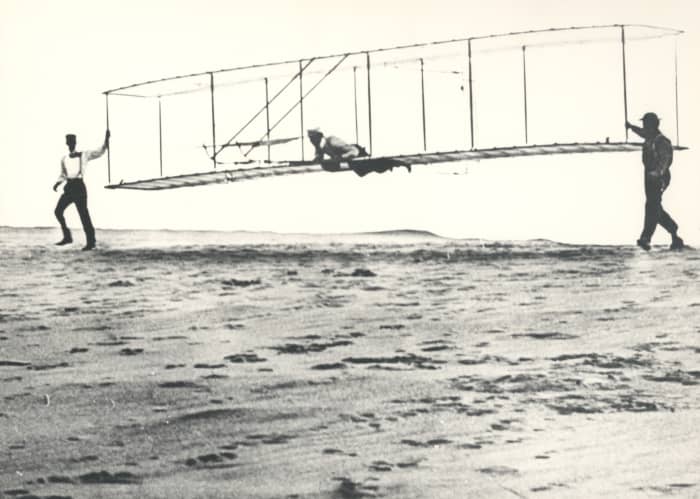 Historic photo of the Wright brothers' third test glider being launched at Kill Devil Hills, North Carolina, on October 10, 1902. Wilbur Wright is at the controls, Orville Wright is at left, and Dan Tate (a local resident and friend of the Wright bro