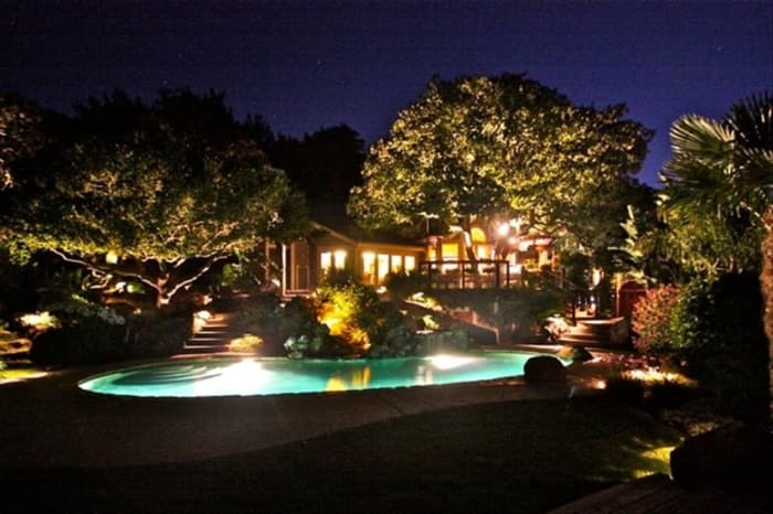 backyard night scape with trees lite from below and foliage surrounding an illuminated swimming pool