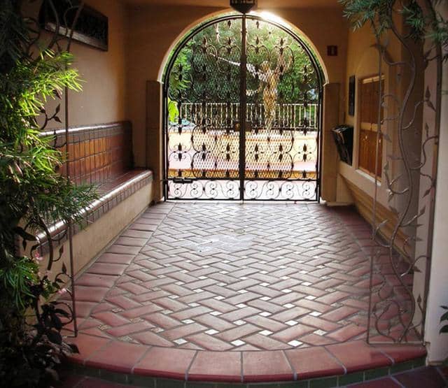 beautiful herringbone terracotta with tan porcelain tile leading to an ornate wrought iron gate and a glimpse of a water fountain with an angel just beyond the covered courtyard