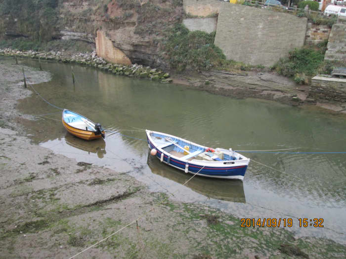 A Yorkshire coble and smaller craft at high tide on the creek, seen from the riverside walk near the small, narrow wooden footbridge