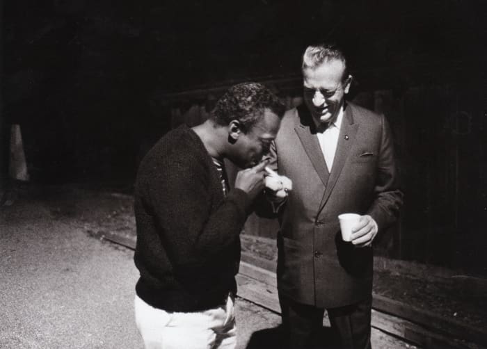 Miles Davis having a cigarette lit by Harry James on the backstage at the Monterey Jazz Festival 1963.