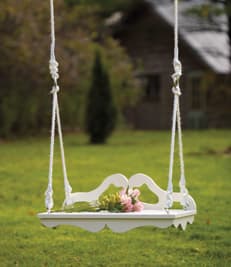 romantic Victorian swing in white with graceful wooden curves with roping and pink roses resting gracefully on the seat