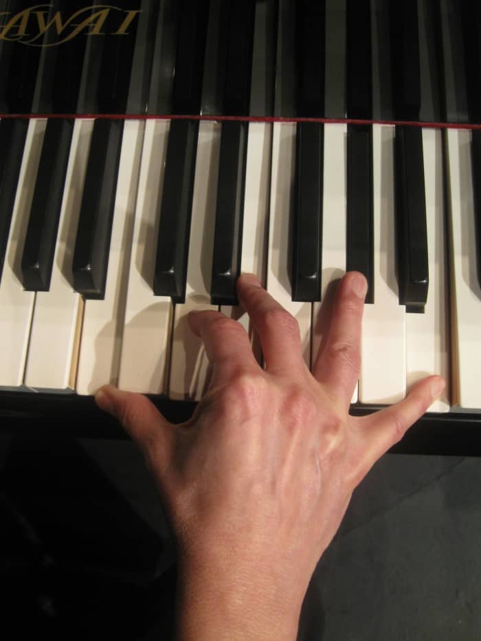 A woman with average sized hands playing a seventh chord on a full size piano. Note the strain of the hand and the awkward position of the fingers.