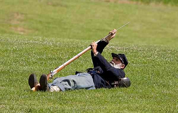 A Living Historian loads his musket from the Prone Stance. He is on his back, balances the firearm between his feet, and is about to ram the cartridge down the barrel