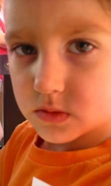 He looks like he is about 8 years old. He has big, dark eyes and a pouting mouth. He is wearing an orange T-shirt. He doesn't look very happy.